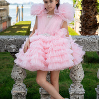 A young girl wearing a pink Aisha tulle flower girl dress with a voluminous skirt and embellished bodice, sitting on a stone bench.