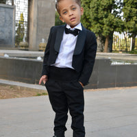 A toddler in a black suit with a white dress shirt and bow tie, standing outdoors.