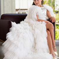 A young girl sitting on a chair, wearing a white tulle flower girl dress with a bodice embellished with white tulle detailing covered with lightweight, sparkly sequins, and a voluminous skirt.