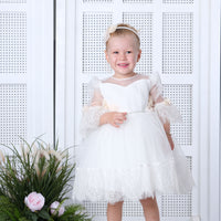 A toddler in a white floral baby dress, standing on a platform with a background of white arches and a floral arrangement.