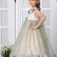 A young girl standing on a platform wearing a green floral girls' dress with a round neckline, and a flare skirt, posing against a backdrop with white arches and decorative flowers.