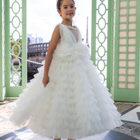A young girl wearing a white tulle flower girl dress with a sleeveless bodice and a full, floor-length skirt, standing in a room with green shutters.