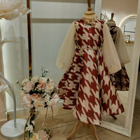 A display mannequin wearing a red and beige geometric patterned dress with a flowy skirt, fitted bodice, and lace detailing. The dress is presented in a retail setting with floral arrangements and reflective mirrors in the background.