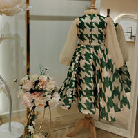 A teenage girl's dress displayed on a mannequin, featuring a geometric pattern with green and beige colors, and a floral arrangement in front.