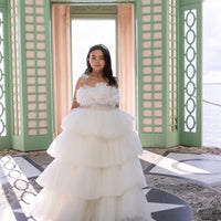 A girl wearing a white tulle flower girl dress with a voluminous skirt and embellished bodice, standing in a doorway.