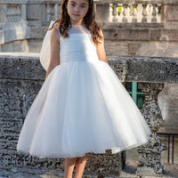 A girl wearing a white tulle flower girl dress with a pleated bodice and a full skirt, standing outdoors.
