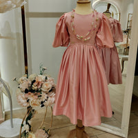 A pink teenage dress with a flowy skirt, short sleeves, and a round neckline, displayed on a mannequin. The dress is adorned with a string of beads, and there are decorative flowers at the bottom.