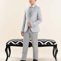 A boy wearing a gray suit with a white shirt and black shoes, standing in front of a black and white bench.