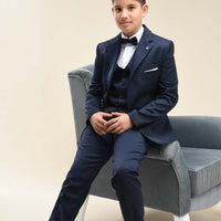 A young boy wearing a navy blue two-piece suit with a jacket and trousers, sitting on a gray chair.