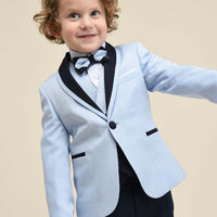A boy wearing a navy suit with a white shirt and tie, standing against a beige background.