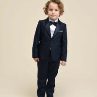 A young boy wearing a black tuxedo with a classic design, including a jacket, trousers, and a white shirt. The tuxedo features a bow tie and a pocket square.