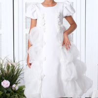 A young girl in a white dress with intricate lace details and a flowing skirt, standing in a room with a white arched doorway and decorative flowers.