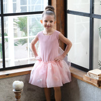 A young girl wearing a pink A-line dress with a bow detail on the waist, standing by a window.