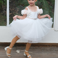 A young girl wearing a white flower girl dress and a full tulle skirt, posing for a photo.
