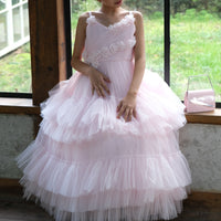 A young girl wearing a pink elegance dress with a fitted bodice, sweetheart neckline, and a full, floor-length skirt with lace detailing. The dress has a back bow and she is sitting by a window.