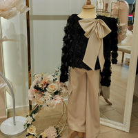 A teenage girl's clothing set displayed on a mannequin, consisting of a black top with a bow detail, beige bottoms, and a floral patterned top. The set is presented in a retail setting with decorative flowers and a reflective mirror behind it.