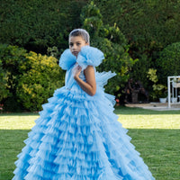 A young girl wearing a sky blue tulle gown with a multilayered skirt and puff sleeves, paired with a sparkling belt and tiara headpiece, standing on grass.