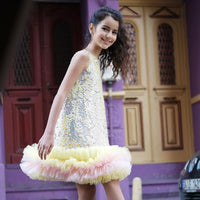 A young girl wearing a sleeveless, knee-length yellow dress with a sequined bodice and a fluffy skirt, standing in front of a purple backdrop.