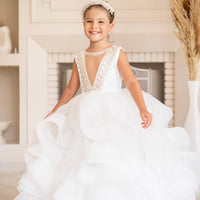 A young girl wearing a white, floral embellished flower girl dress with a V-neckline and sleeveless design, standing in a room with a fireplace.