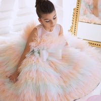 A young girl wearing a pink and multicolored tulle dress with a bodice featuring embroidery, sitting in a thoughtful pose with a smile on her face.