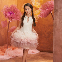 A girl wearing a pink dress with a flowy skirt and floral embellishments, standing in front of a orange and pink backdrop with artificial flowers.