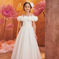 A girl wearing an off-white, floral patterned dress with short puffed sleeves and a maxi skirt, standing in a room with a floral backdrop.