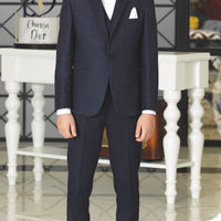 A boy wearing a blue suit with a white shirt and tie, standing in a room with white furniture.