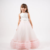 A young girl wearing a sleeveless pink tulle dress with train, featuring pearl and bead detailing, standing against a white background.