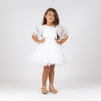 A young girl standing against a white background, wearing a white floral dress with three-quarter sleeves and a cinched waist, paired with ballet shoes.