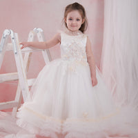 A young girl wearing a white sleeveless dress with a rich satin bodice, delicate two-colored lace with crystal cut beads, and a tulle skirt trimmed with ruffled tulle. The dress features a V-cut back and a back zipper.