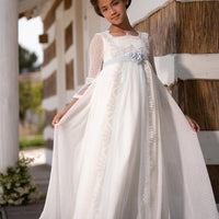A young girl wearing a traditional Spanish long white dress with intricate lace detailing, posing on a wooden porch with a rustic backdrop.