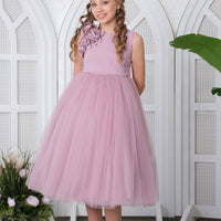 A girl in a purple tulle dress with floral details on the bodice and a bow at the back, standing in a room with a white brick wall and a potted plant in the background.