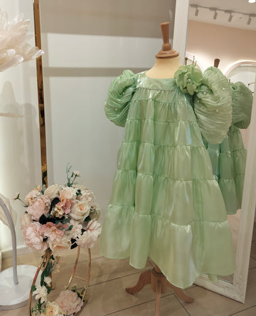 A green, tiered, sleeveless girls' dress on a mannequin, with a flower arrangement in the background.