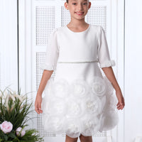 A young girl standing in a beautifully decorated room wearing a white flower girl dress with lace and floral details, and a bow on the bodice.