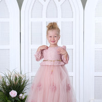 A baby girl wearing a pink floral baby dress, standing on a platform with white arched doors in the background.