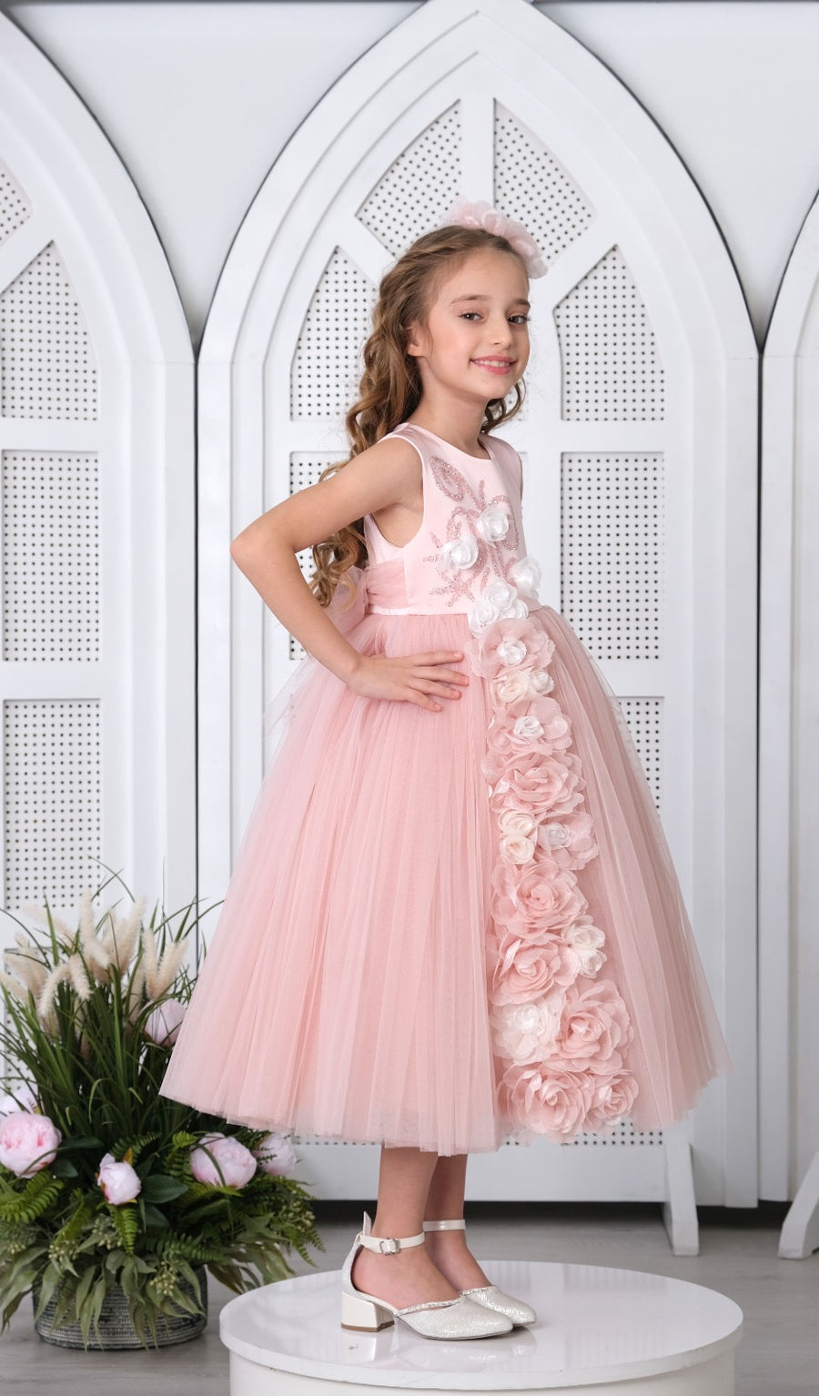 A young girl standing on a platform wearing a pink floral dress with a flowy skirt, paired with white shoes and a flower crown.